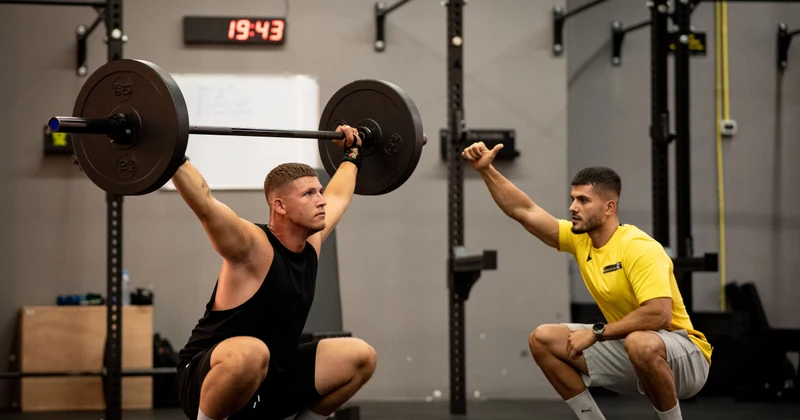 Coach supervising a barbell lift in a strength-training gym.